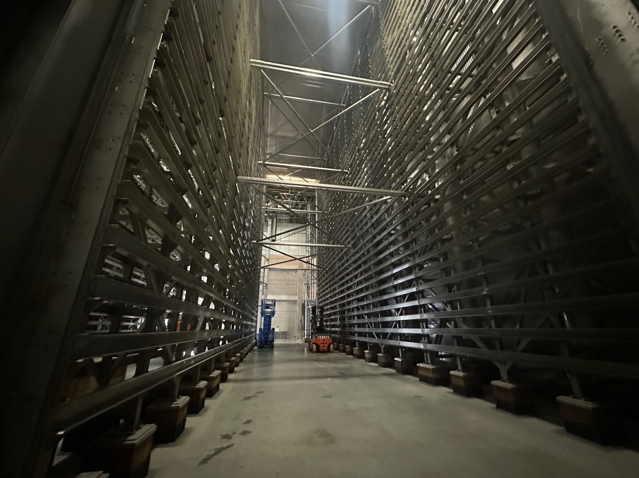 Wnętrze magazynu z regałami metalowymi, widok na wózek widłowy i podnośnik koszowy w oddali.