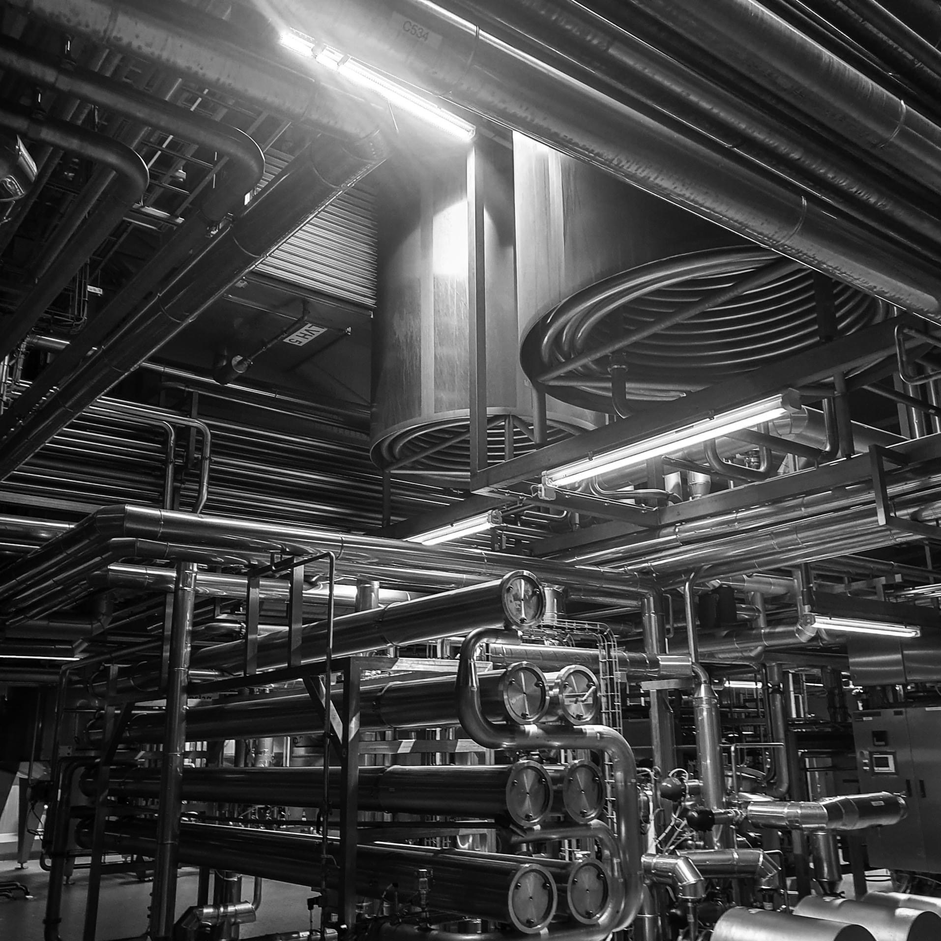 Czarno-białe zdjęcie przedstawiające skomplikowany system rur przemysłowych z zaworami i manometrami, oświetlonych sztucznym światłem, tworzących labirynt instalacji.