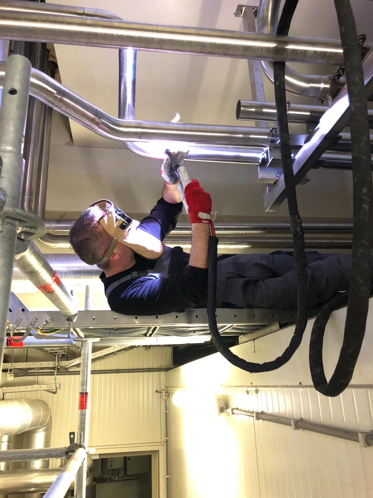 Pracownik w masce spawalniczej, leżąc na rusztowaniu, łączy rury ze stali nierdzewnej wewnątrz budynku przemysłowego, używając metody spawania TIG.