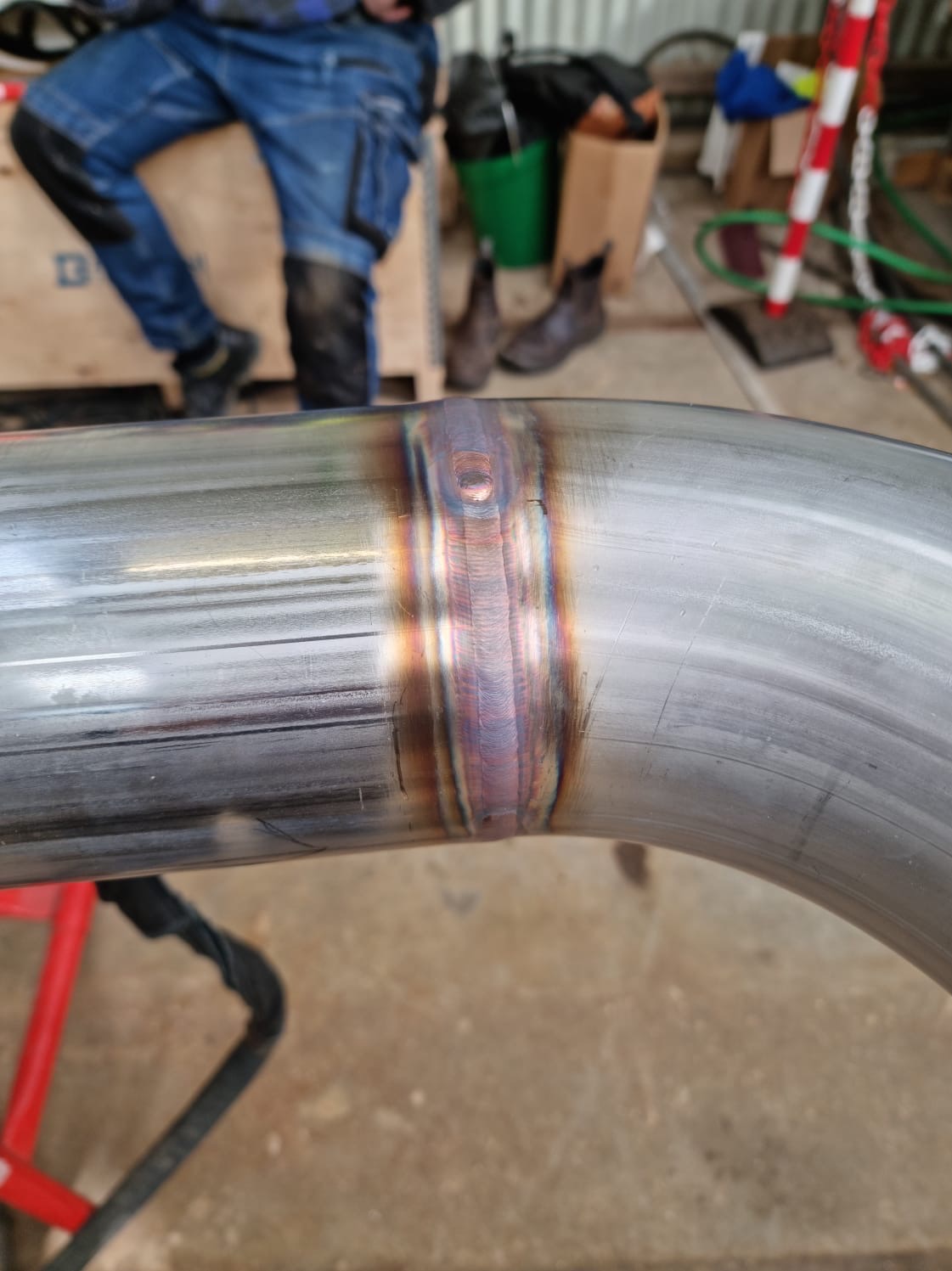 Spoina na rurze ze stali nierdzewnej, widoczne charakterystyczne przebarwienia po spawaniu, w tle robocze ubranie i obuwie osoby wykonującej prace.