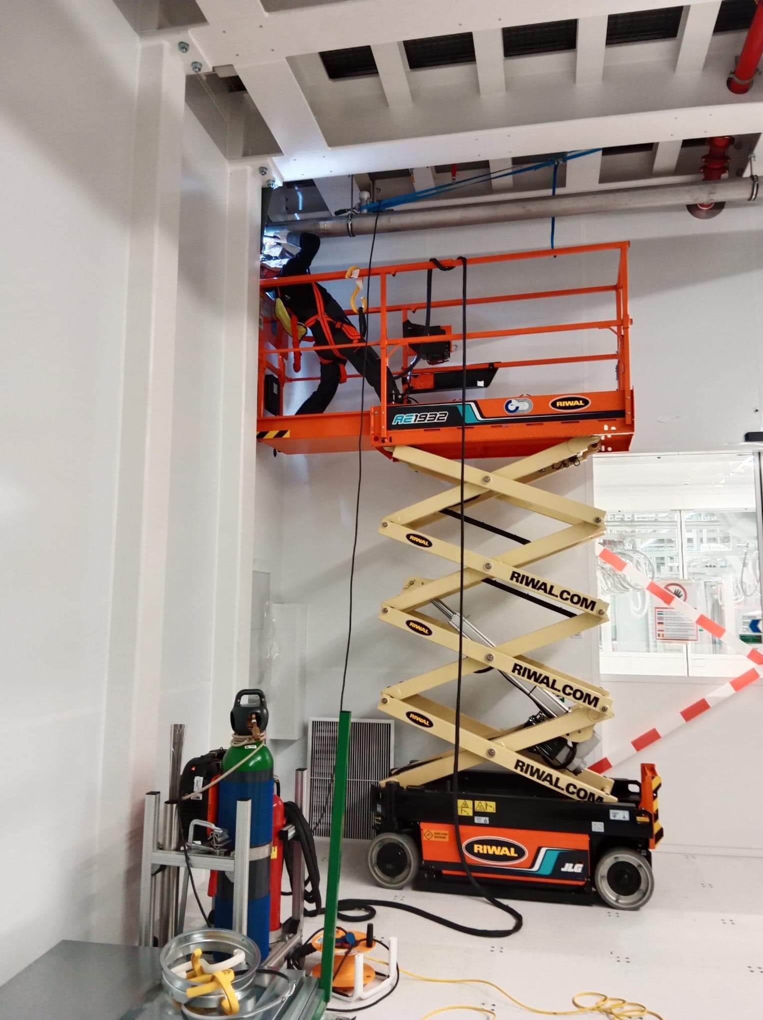 Pracownik w kombinezonie i masce spawalniczej na podnośniku nożycowym, wykonujący prace spawalnicze przy suficie w pomieszczeniu o białych ścianach. Widoczny wózek z butlami gazowymi i sprzętem...