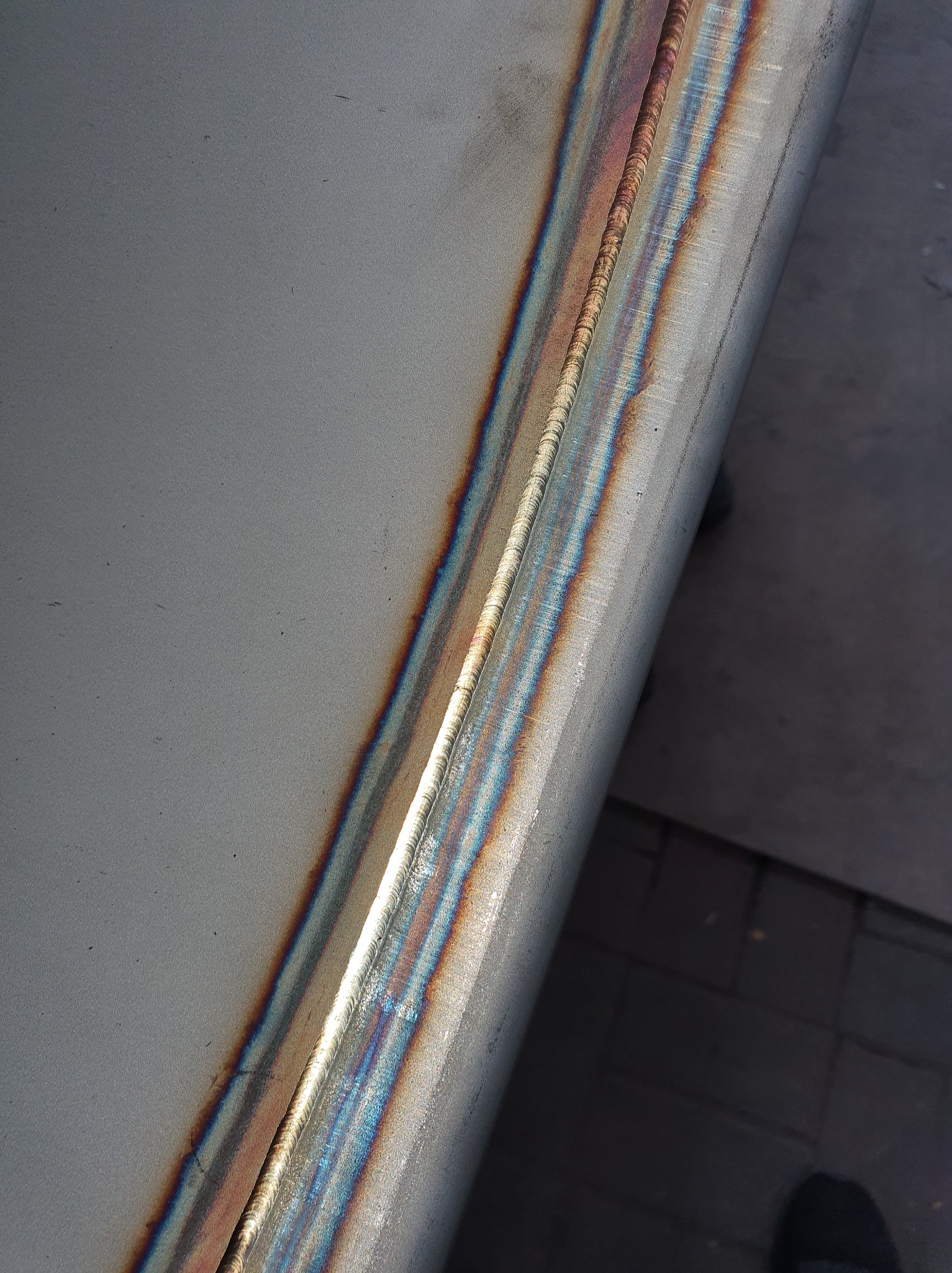 Szlifowana powierzchnia metalowa z widocznym spawem o opalizujących barwach termicznych, położona na szarym tle.