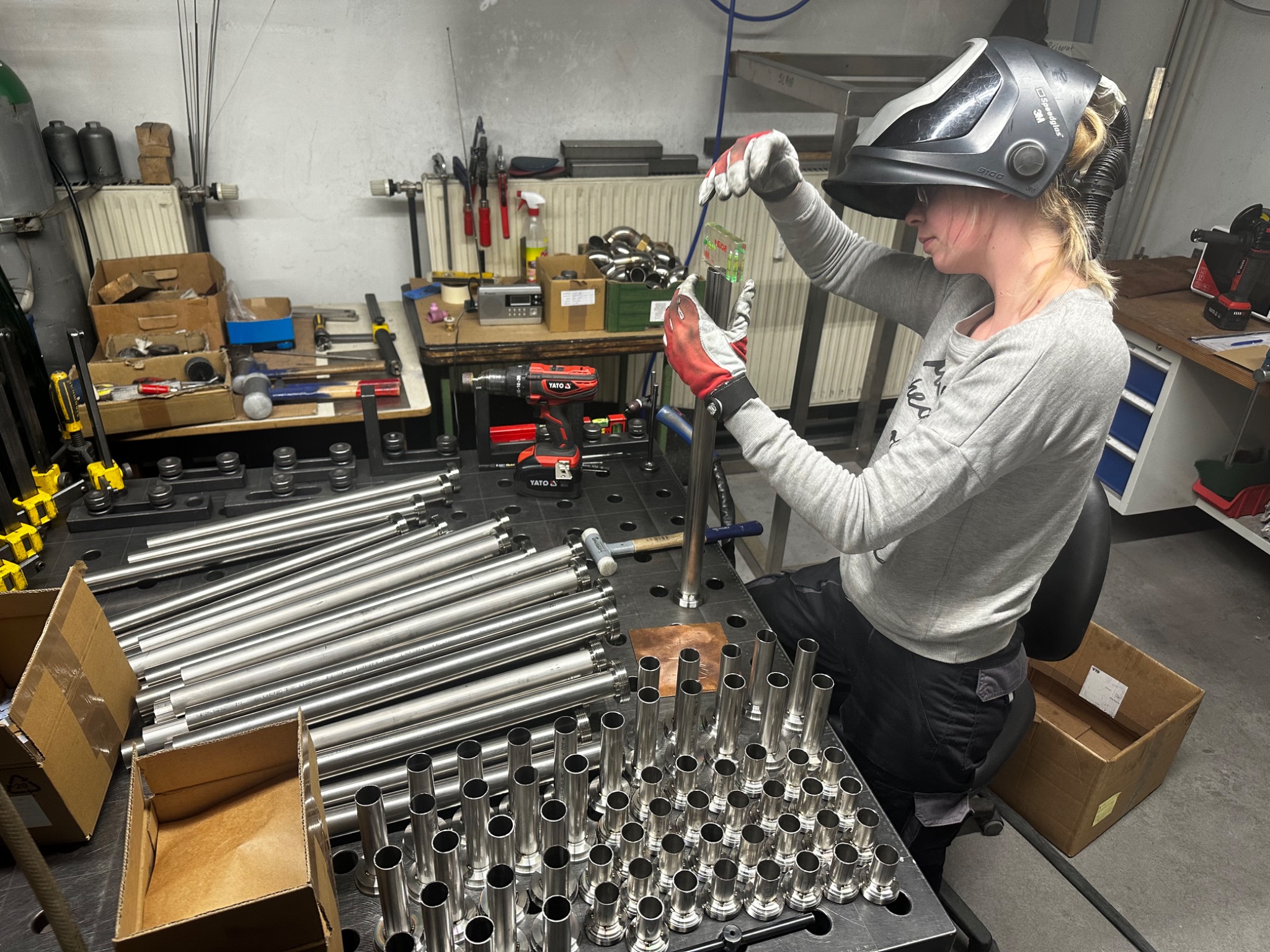 Pracownik w Elblągu w masce spawalniczej przygotowuje element metalowy do obróbki, używając poziomicy na warsztacie z narzędziami i rurami.