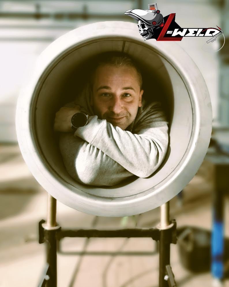 Mężczyzna uśmiecha się, siedząc wewnątrz dużej, spawanej rury na metalowym stojaku, z logo firmy w rogu.