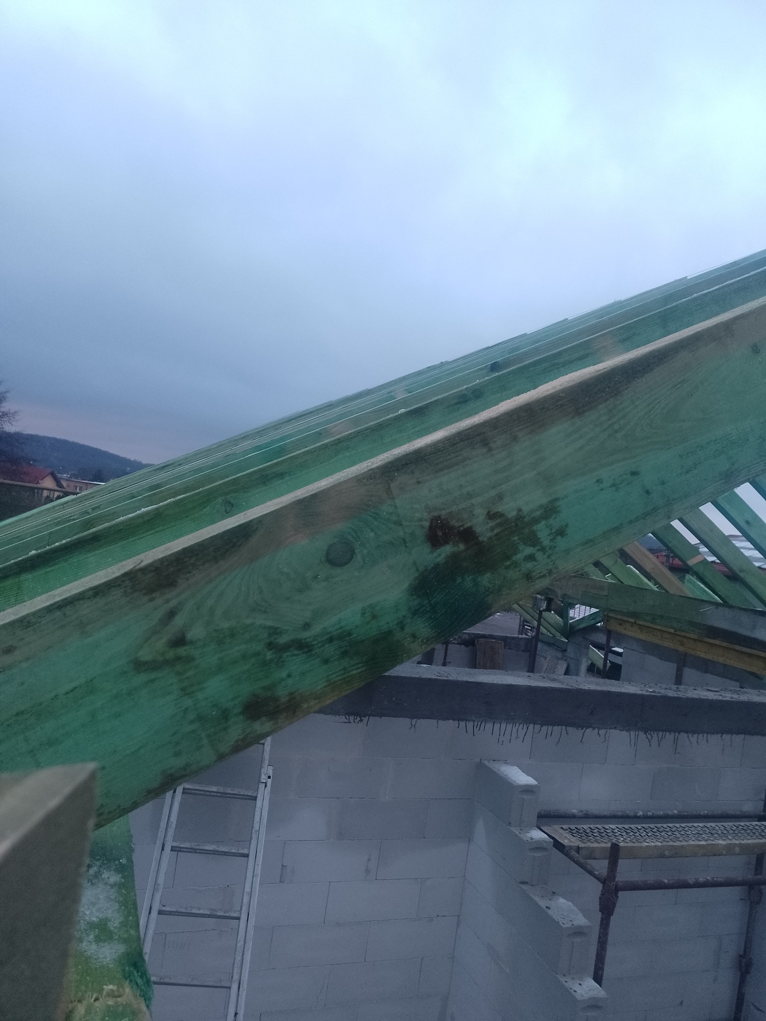 Wiązary dachowe zaimpregnowane na zielono, widoczne na tle murowanych ścian z betonu komórkowego i drabiny, w tle zabudowa jednorodzinna