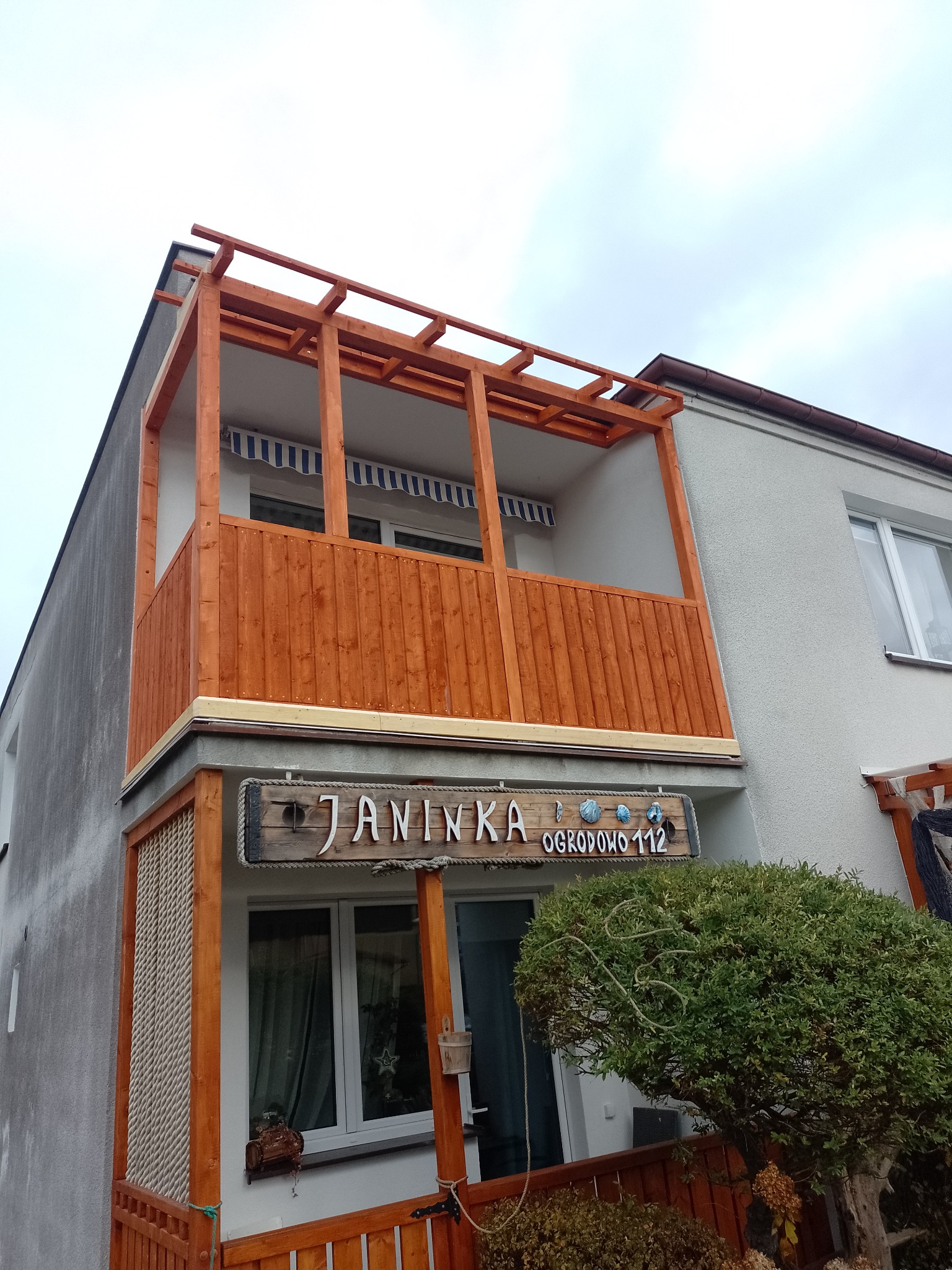 Nowo wybudowany, drewniany balkon z zadaszeniem nad wejściem do budynku z tabliczką adresową 'JANINKA OGRODOWO 112'.