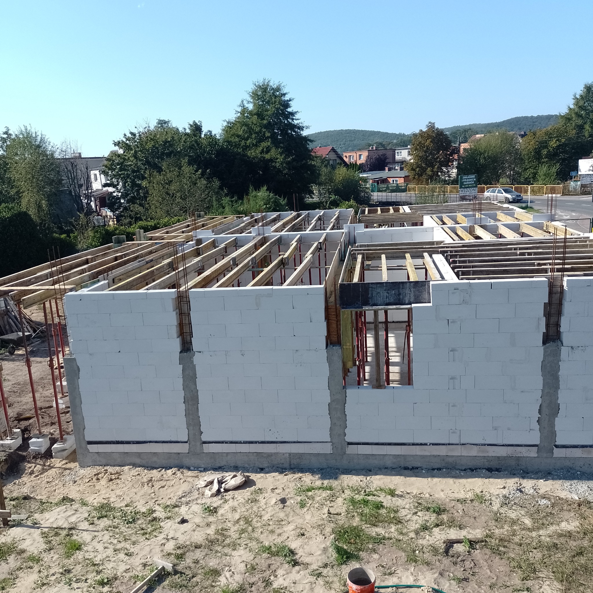Wznoszenie ścian parteru z białych bloczków, widoczne zbrojenia i drewniane elementy konstrukcji stropu, w tle zabudowa jednorodzinna i zieleń.