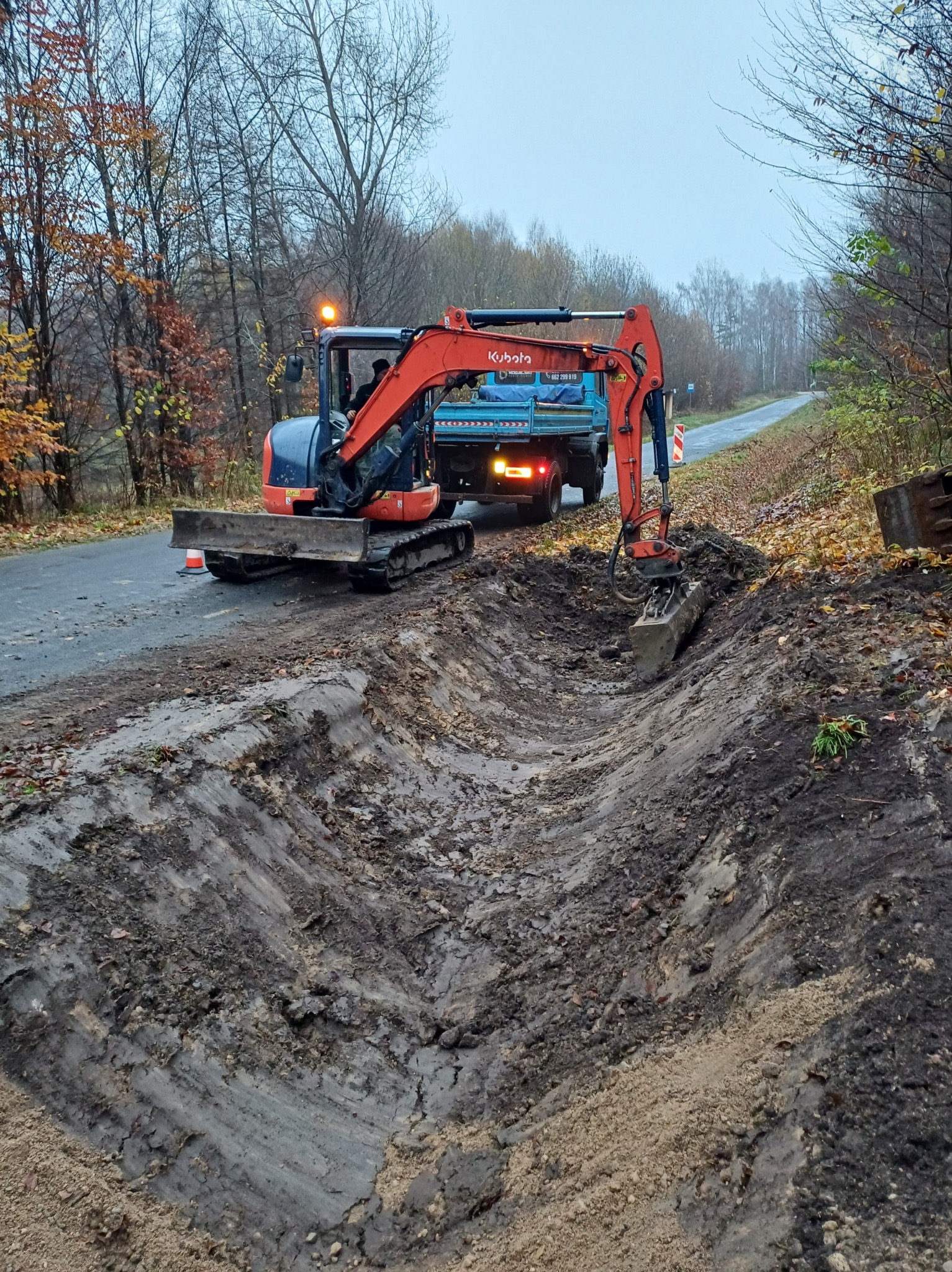 Pomarańczowy minikoparka Kubota na poboczu asfaltowej drogi, wykopany rów z błotnistą ziemią, w tle ciężarówka i jesienny krajobraz drzew