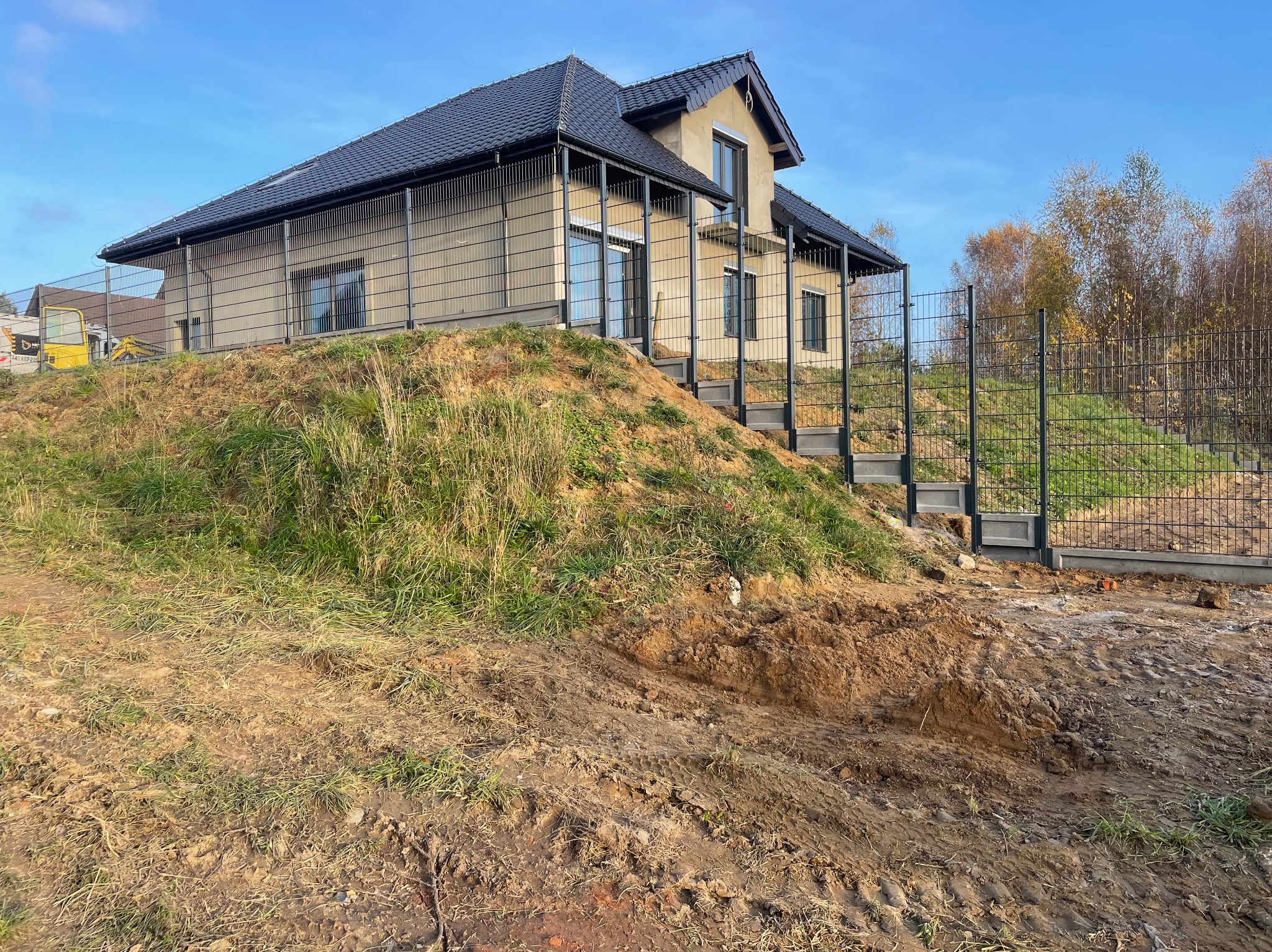 Nowoczesny dom jednorodzinny na wzniesieniu, otoczony ogrodzeniem z siatki; widoczne ślady prac ziemnych i żółta koparka w tle.