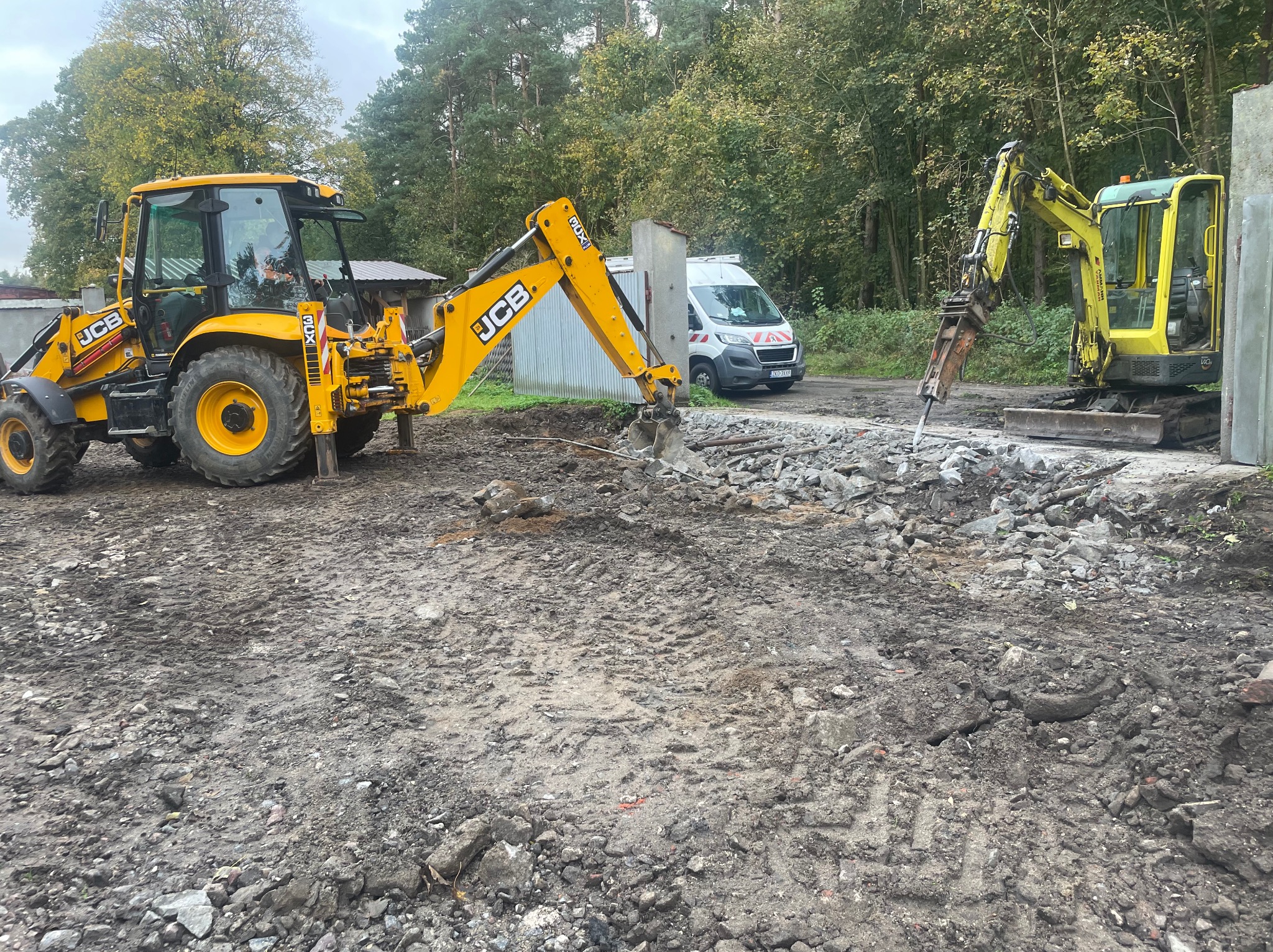 Żółta koparko-ładowarka JCB z przodu i mniejsza żółta koparka z wiertłem hydraulicznym rozbijająca beton na placu budowy z gruzem i śladami opon, na tle drzewa i białego samochodu dostawczego.