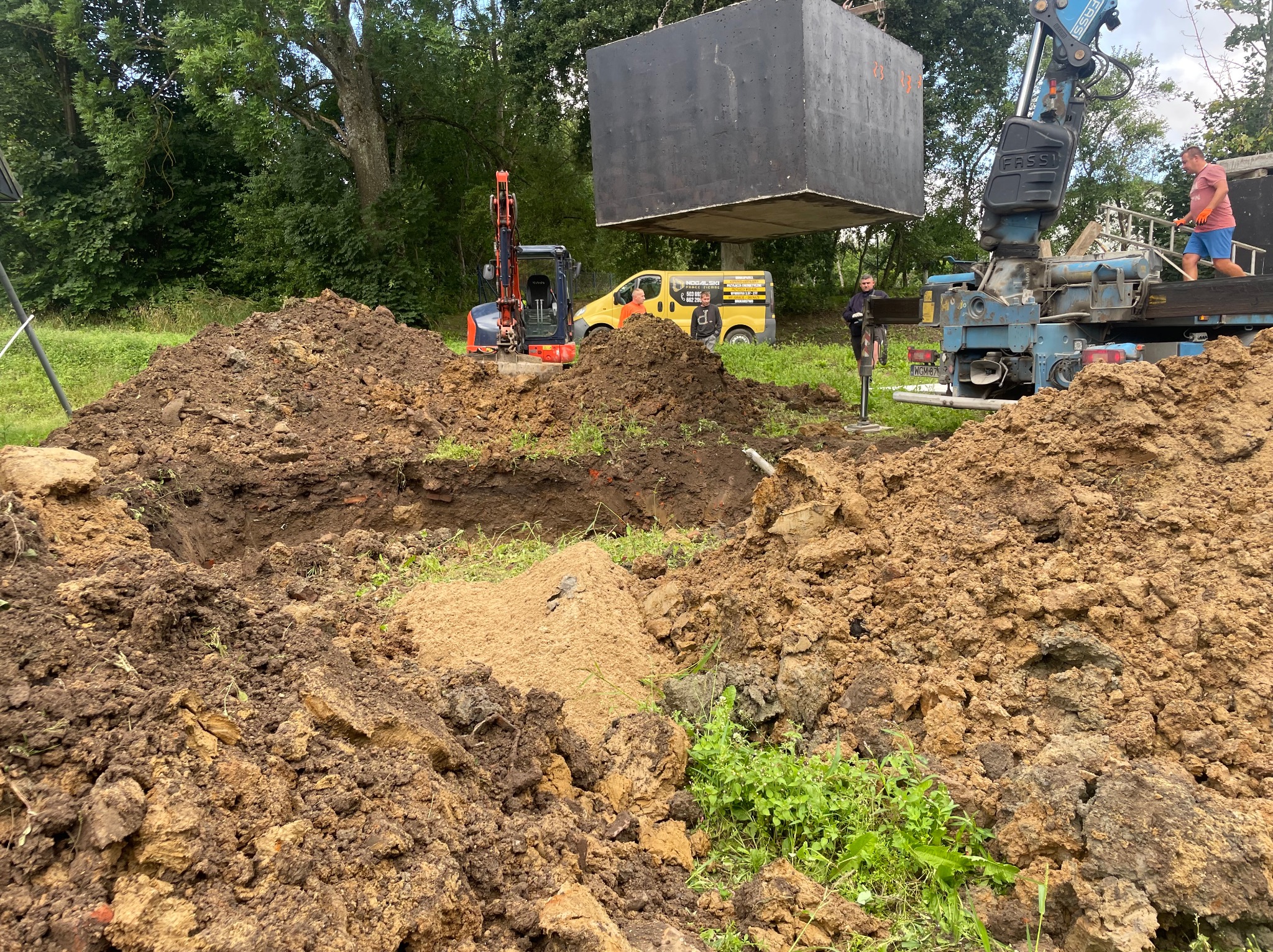 Czarny, betonowy blok podnoszony przez dźwig na placu budowy, z wykopem w ziemi i minikoparką w tle.