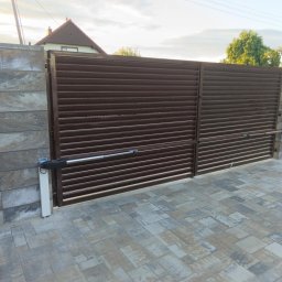 Spaw-Art Mateusz Godlewski - Automatyczna, brązowa brama wjazdowa z poziomych profili, zamontowana na kamiennych słupkach. Kostka brukowa na podjeździe. Widoczne siłowniki bramy.