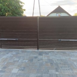 Spaw-Art Mateusz Godlewski - Automatyczna brama wjazdowa, brązowa, poziome lamele. Kostka brukowa, słupki z kamienia. Napęd bramy widoczny. W tle dom i zieleń. Ujęcie z poziomu gruntu.