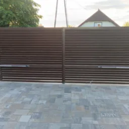 Automatyczna brama wjazdowa, brązowa, poziome lamele. Kostka brukowa, słupki z kamienia. Napęd bramy widoczny. W tle dom i zieleń. Ujęcie z poziomu gruntu.