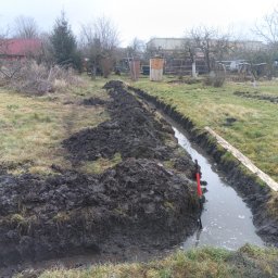 Spaw-Art Mateusz Godlewski - Wykopany rów w ziemi, częściowo wypełniony wodą, z pryzmą ziemi obok. W tle widoczne drzewa i zabudowania. Widoczna łopata z czerwoną rączką.
