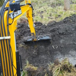 Spaw-Art Mateusz Godlewski - Żółta minikoparka w trakcie prac ziemnych, z bliska widoczny lemiesz zagłębiający się w mokrą glebę. Widoczne zielone tło z drzewami i trawą.