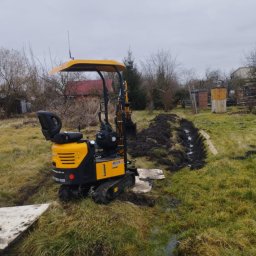 Spaw-Art Mateusz Godlewski - Żółta minikoparka na trawiastym terenie, wykopany rów w tle, pochmurne niebo. Widoczny panel z numerem telefonu. Praca w toku.