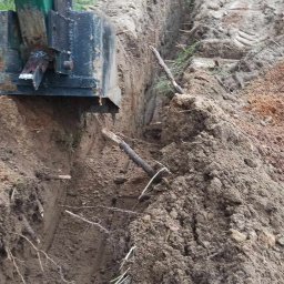 Spaw-Art Mateusz Godlewski - Wykopany wąski rów w ziemi z fragmentem zielonej minikoparki. W tle ogrodzenie z siatki i drzewa.