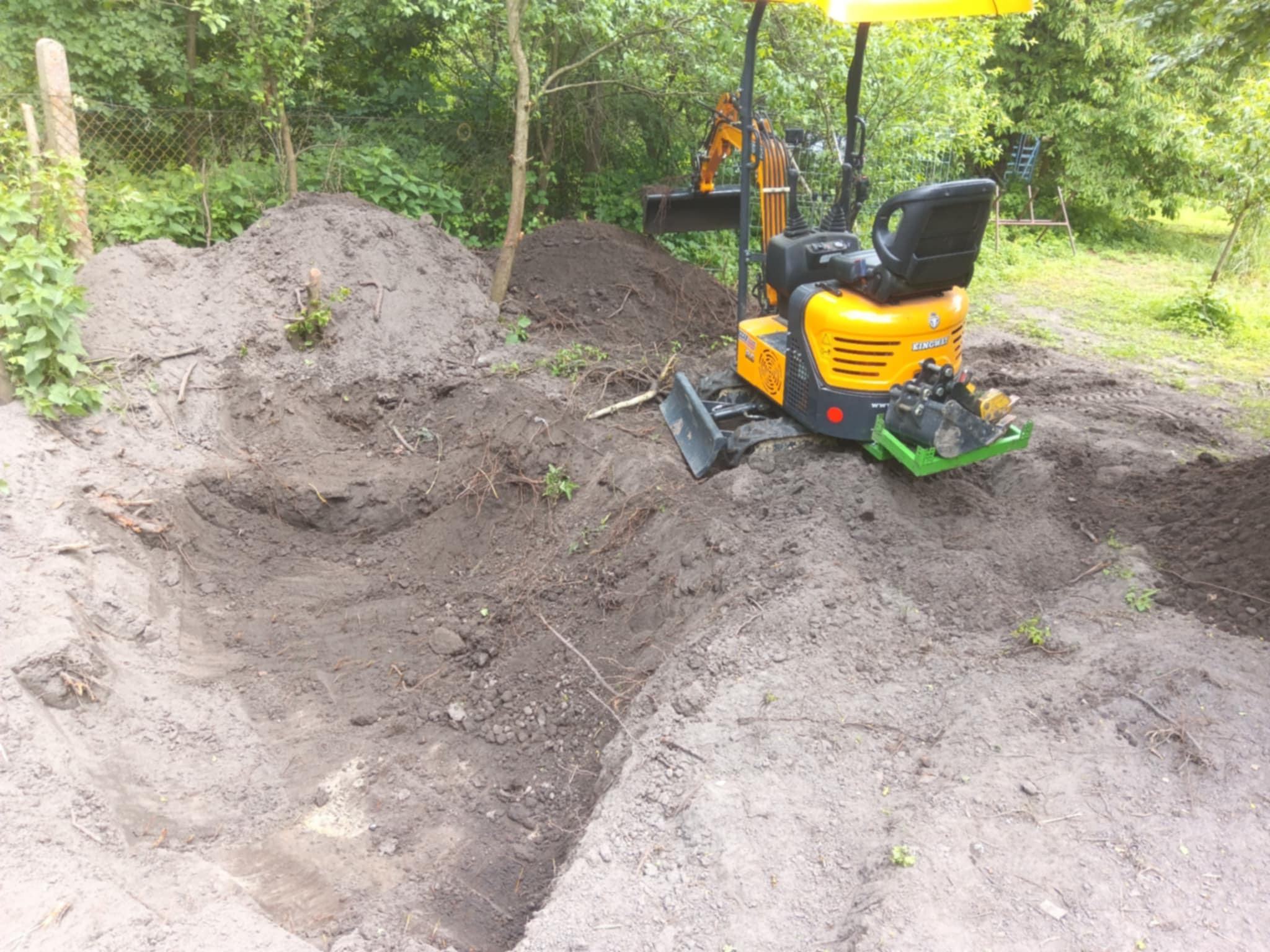 Żółta minikoparka na ziemi, obok wykopany dół. W tle zieleń drzew. Widoczny etap prac ziemnych, przygotowanie terenu pod inwestycję.