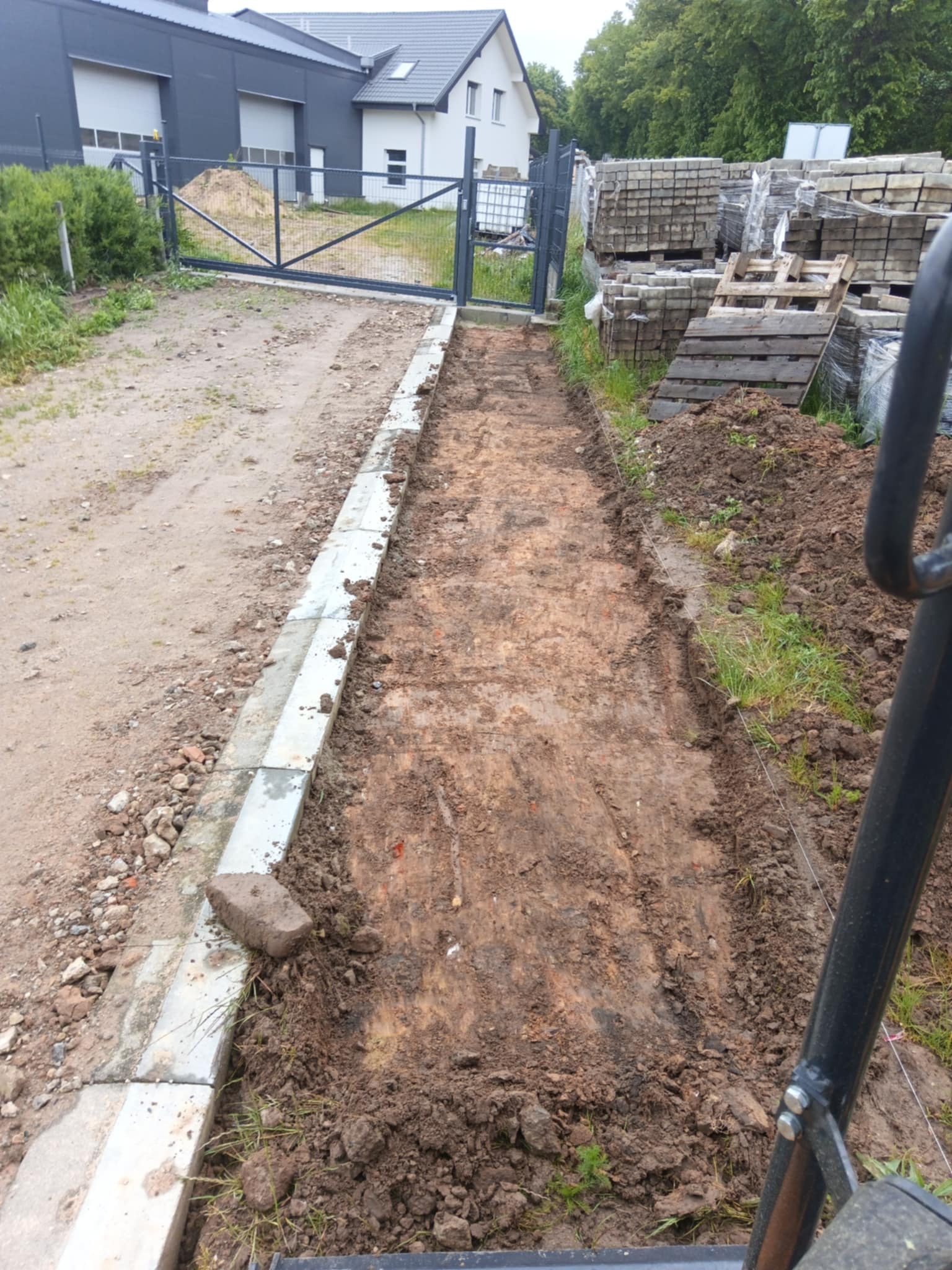 Przygotowanie terenu pod ścieżkę: wykopana ziemia, obrzeża z kostki, w tle brama wjazdowa i budynek. Widoczne palety z kostką brukową.