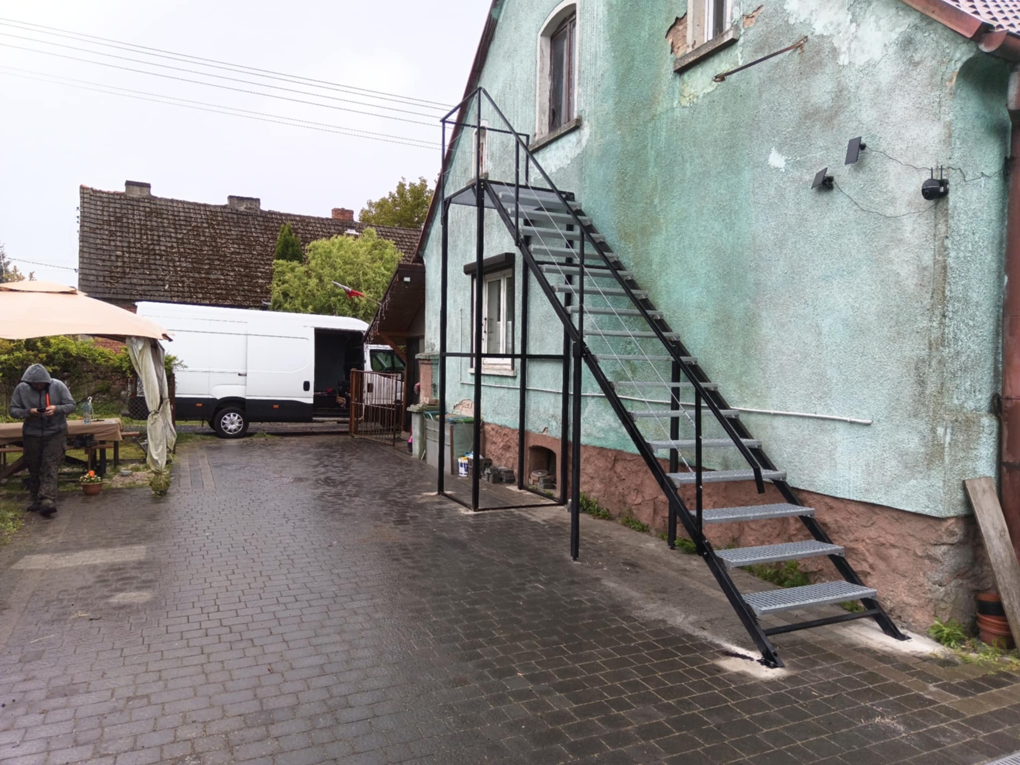 Zewnętrzne schody metalowe z kratowymi stopniami, prowadzące na piętro budynku z zielonkawą elewacją. Widoczne kamery przemysłowe. Na podwórku zaparkowany biały van.