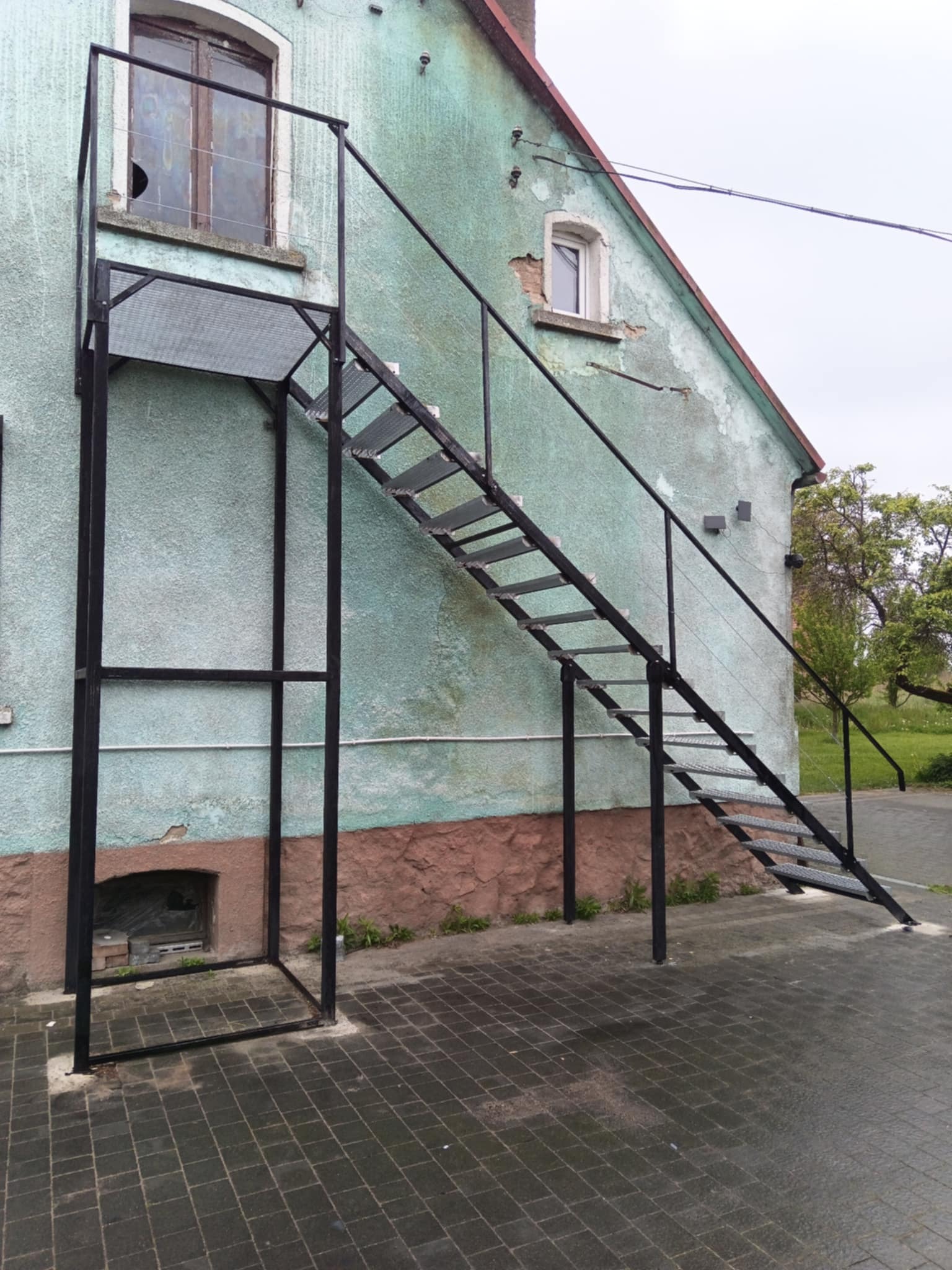Zewnętrzne schody metalowe prowadzące do okna na piętrze budynku z zieloną, łuszczącą się elewacją i brązową podmurówką. Metalowa konstrukcja pomalowana na czarno.