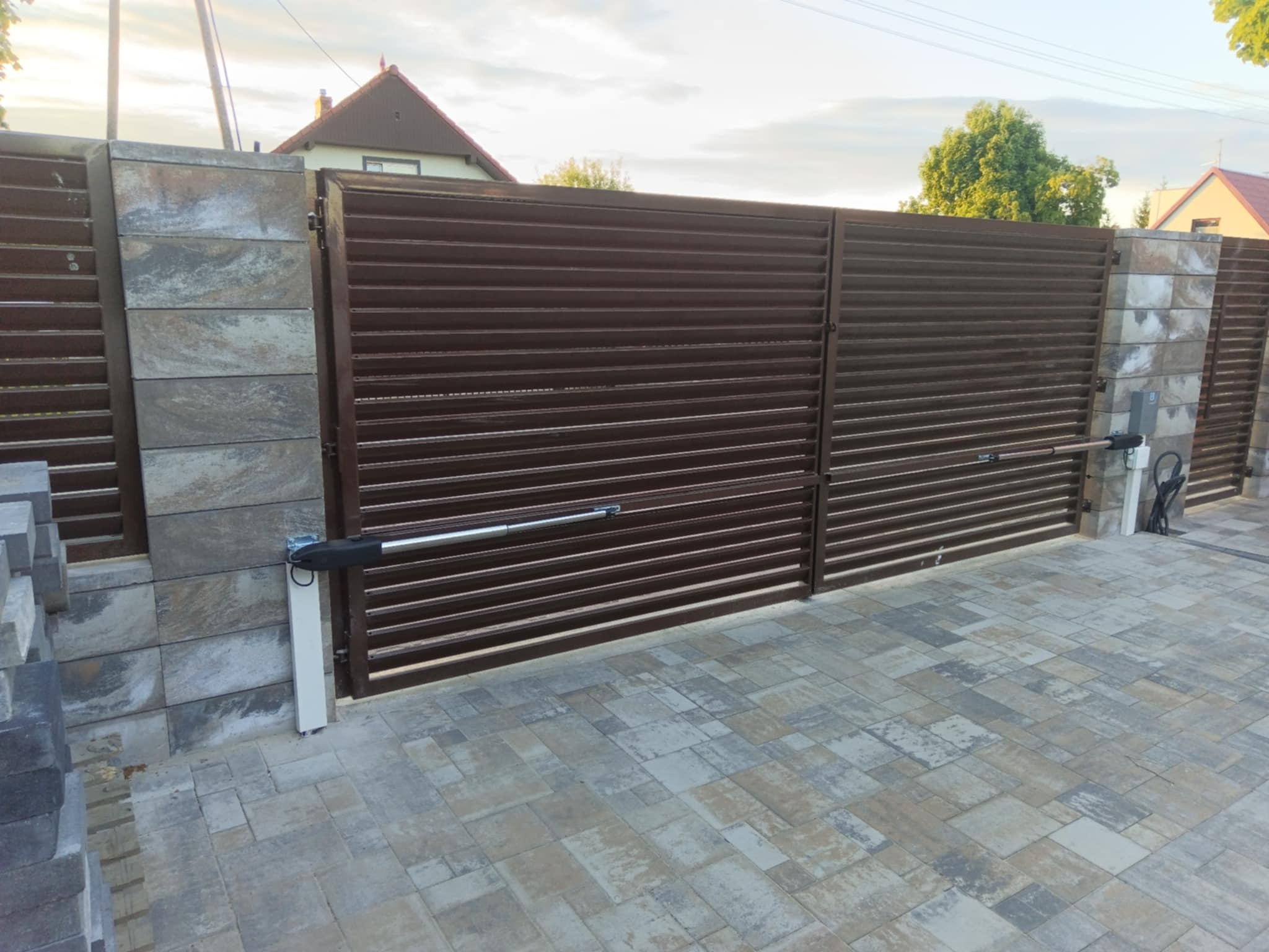 Automatyczna, brązowa brama wjazdowa z poziomych profili, zamontowana na kamiennych słupkach. Kostka brukowa na podjeździe. Widoczne siłowniki bramy.