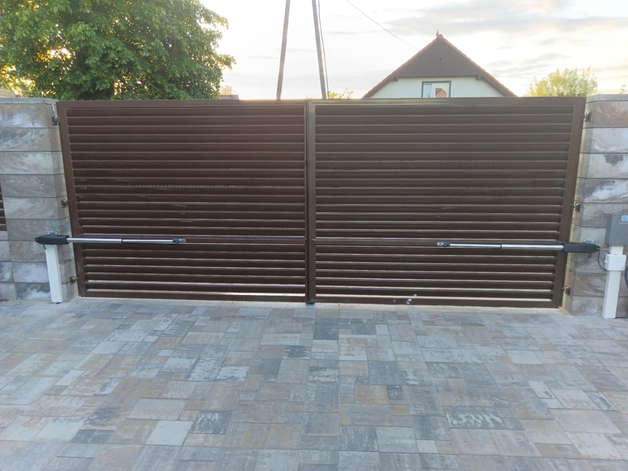 Automatyczna brama wjazdowa, brązowa, poziome lamele. Kostka brukowa, słupki z kamienia. Napęd bramy widoczny. W tle dom i zieleń. Ujęcie z poziomu gruntu.