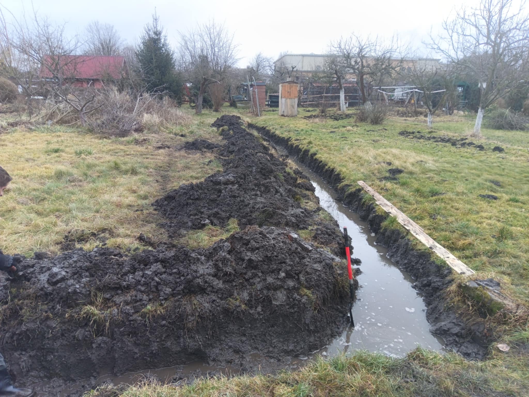 Wykopany rów w ziemi, częściowo wypełniony wodą, z pryzmą ziemi obok. W tle widoczne drzewa i zabudowania. Widoczna łopata z czerwoną rączką.