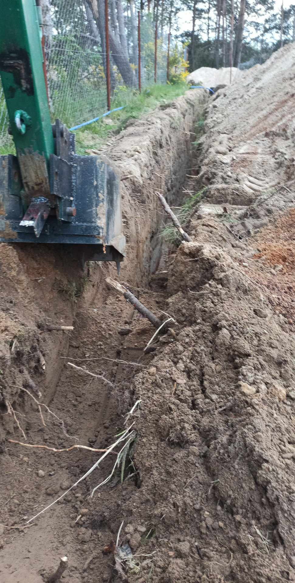 Wykopany wąski rów w ziemi z fragmentem zielonej minikoparki. W tle ogrodzenie z siatki i drzewa.