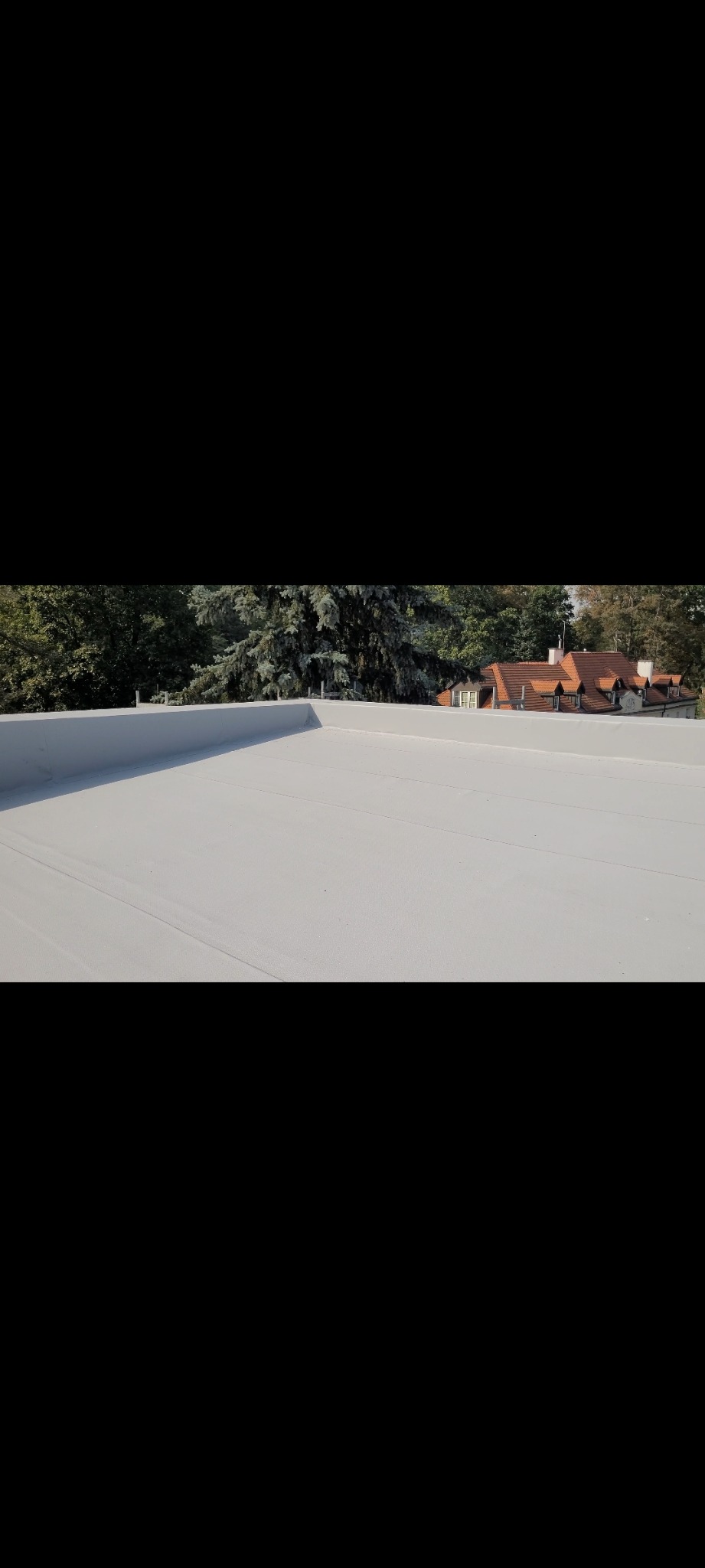 Nowy, jasnoszary dach płaski z widocznymi liniami łączeń, w tle drzewa i fragment domu z czerwonym dachem.