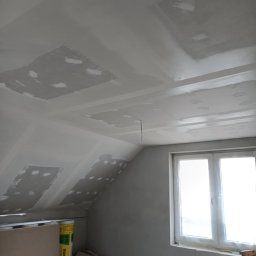 Maj-bud - Skośny sufit poddasza z widocznymi łatami świeżo nałożonej gładzi szpachlowej, przygotowany do malowania, obok okno i materiały budowlane.
