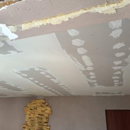 Maj-bud - Nierówna powierzchnia sufitu w trakcie naprawy z widocznymi łatami gipsowymi i odsłoniętym fragmentem starej konstrukcji, z fragmentem ściany z dekoracyjną okładziną kamienną w dolnej części kadru.