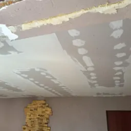 Nierówna powierzchnia sufitu w trakcie naprawy z widocznymi łatami gipsowymi i odsłoniętym fragmentem starej konstrukcji, z fragmentem ściany z dekoracyjną okładziną kamienną w dolnej części kadru.