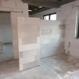 Wnętrze w trakcie budowy, ściany z białych bloczków, widoczne drewniane elementy konstrukcji dachu, surowy beton na podłodze.