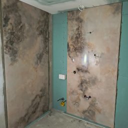 Maj-bud - Wnętrze łazienki w trakcie remontu, z zamontowanymi płytami gipsowo-kartonowymi i częściowo położonymi płytkami imitującymi marmur na ścianie, widoczne instalacje hydrauliczne i elektryczne.