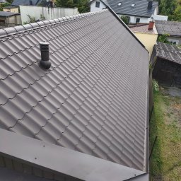 Maj-bud - Nowy, brązowy dach z blachodachówki modułowej z kominkiem wentylacyjnym na tle zabudowy miejskiej i zieleni. Widok z góry, ukośny kadr.