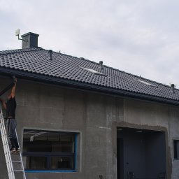 Maj-bud - Monter na drabinie instaluje rynnę na tle surowej elewacji domu z ciemnoszarym dachem. Widoczne okna z niebieską taśmą zabezpieczającą.