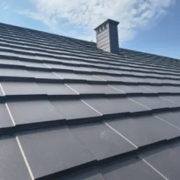 Szary, metalowy dach z kominem na tle błękitnego nieba z chmurami. Widok z dołu ukazuje regularne ułożenie dachówek i estetyczne wykończenie komina.