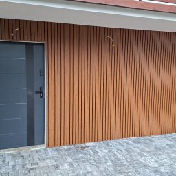 Maj-bud - Elewacja budynku wykończona drewnopodobnymi panelami pionowymi, z szarą, nowoczesną stolarką drzwiową i kostką brukową na podłożu. Detal architektoniczny.
