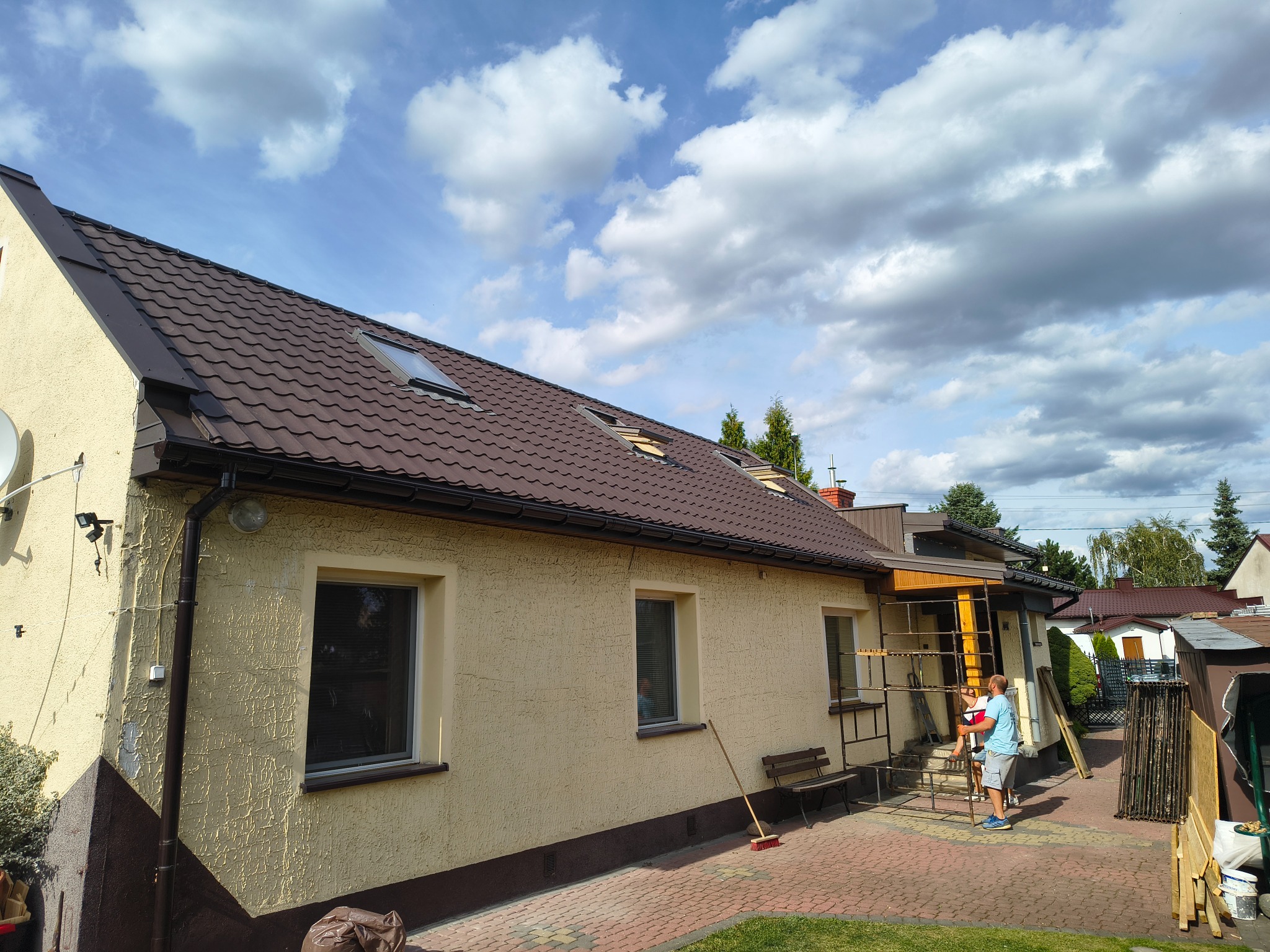 Dom z brązowym dachem i beżową elewacją, widoczne rusztowania i osoby. Ujęcie w słoneczny dzień z częściowo zachmurzonym niebem.