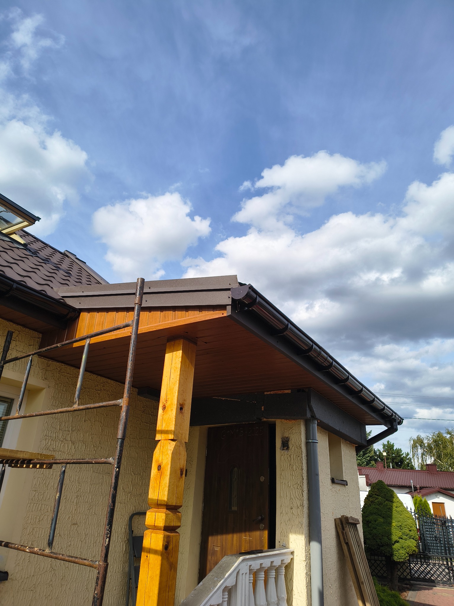 Montaż rynien w Radomiu: Brązowe rynny zamontowane na drewnianej podbitce dachu. Widoczne rusztowanie i fragment dachu z oknem połaciowym na tle błękitnego nieba.