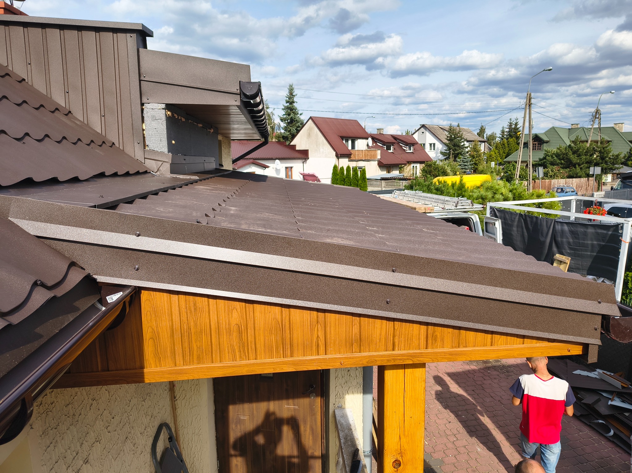 Brązowy dach z blachy trapezowej z rynną, widok z góry, fragment elewacji z drewnianą boazerią, w tle domy i błękitne niebo z chmurami.
