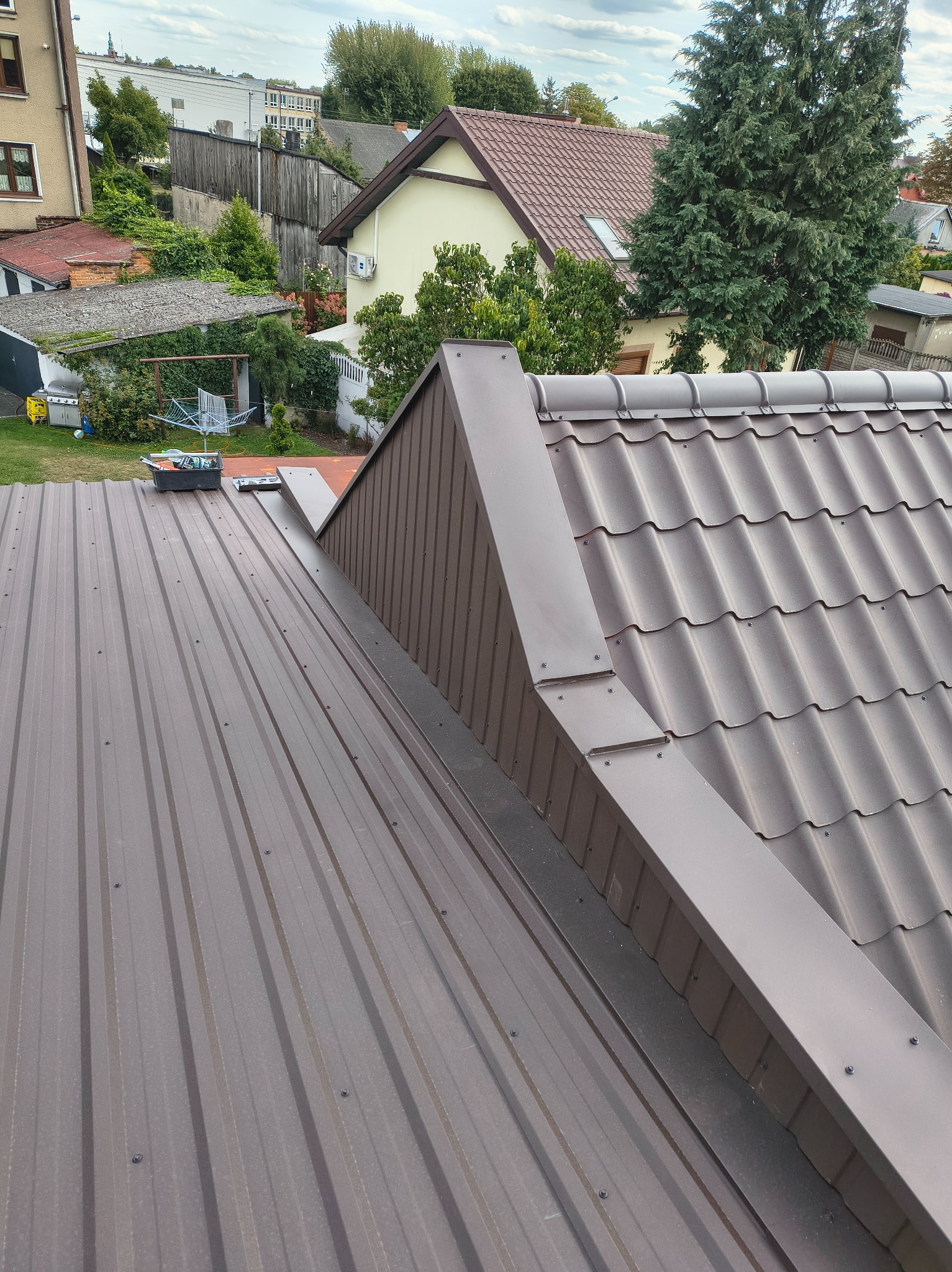 Nowy, brązowy dach z blachy trapezowej i dachówki modułowej, z widocznymi obróbkami blacharskimi, na tle zabudowy jednorodzinnej i zieleni.