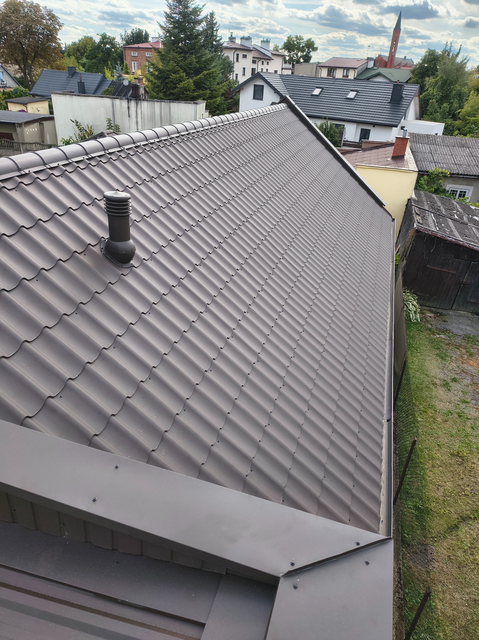 Nowy, brązowy dach z blachodachówki modułowej z kominkiem wentylacyjnym na tle zabudowy miejskiej i zieleni. Widok z góry, ukośny kadr.