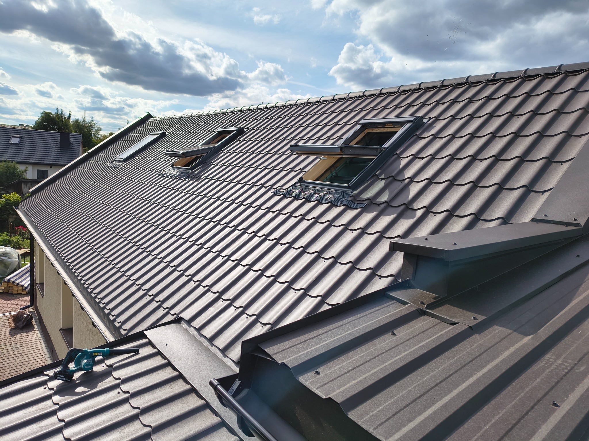 Nowy, brązowy dach z dachówki modułowej i oknami dachowymi, widok z góry, na dachu leży dmuchawa akumulatorowa, obróbki blacharskie w kolorze dachu.