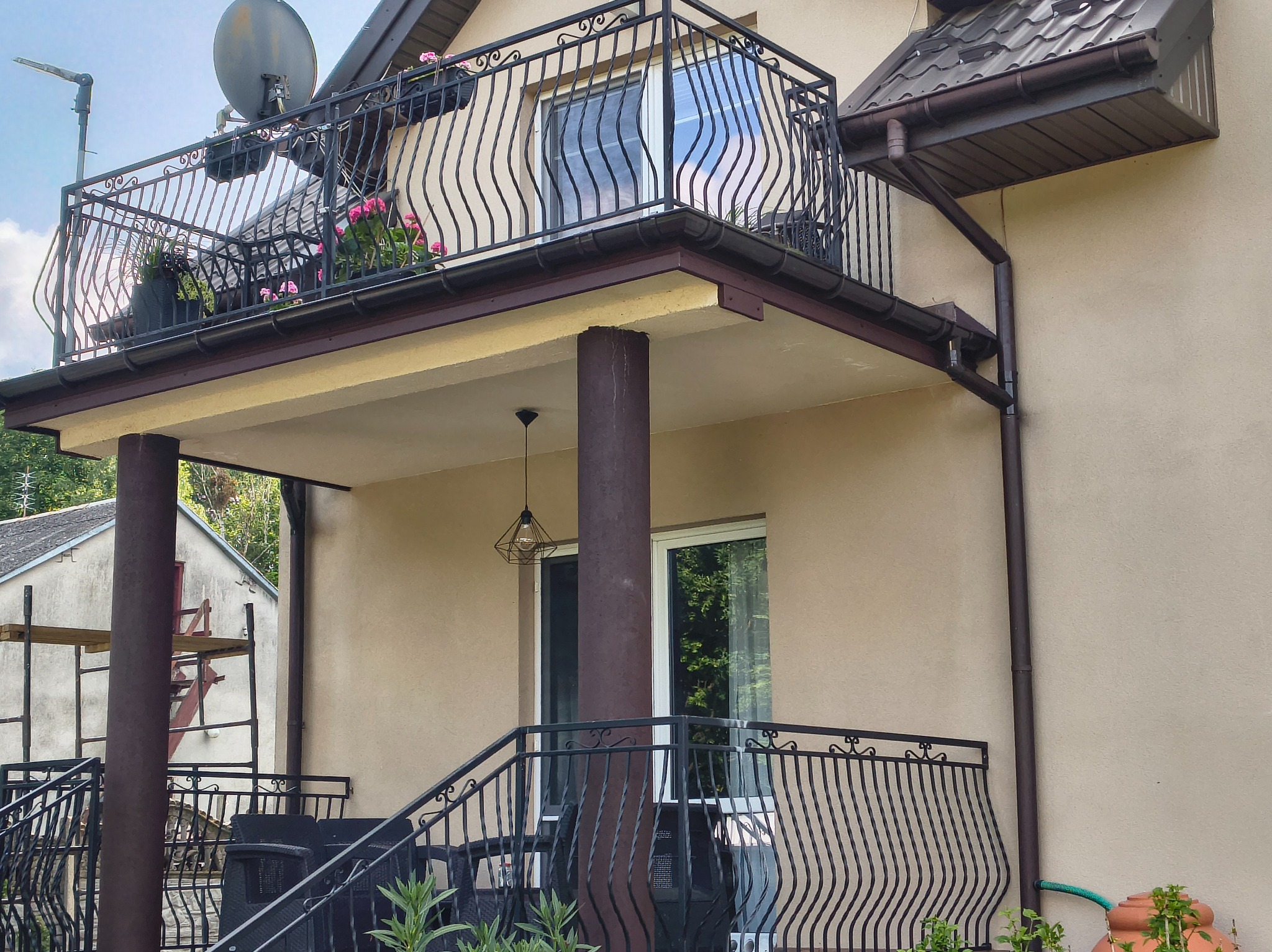 Elewacja domu z balkonem i kutymi balustradami. Widoczne rynny, dachówka i dekoracyjne elementy. W tle rusztowanie przy sąsiednim budynku.