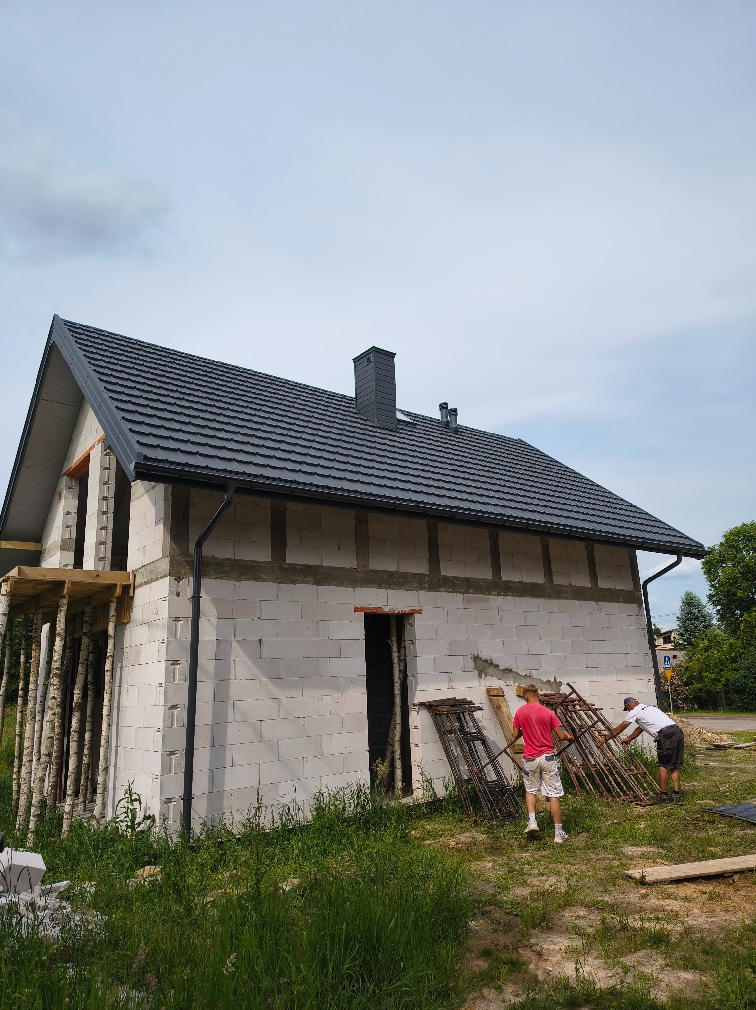 Dom w trakcie budowy z czarnym dachem i rynnami. Dwóch mężczyzn pracuje przy rusztowaniu. Widoczne białe bloczki i drewniane podpory balkonu.
