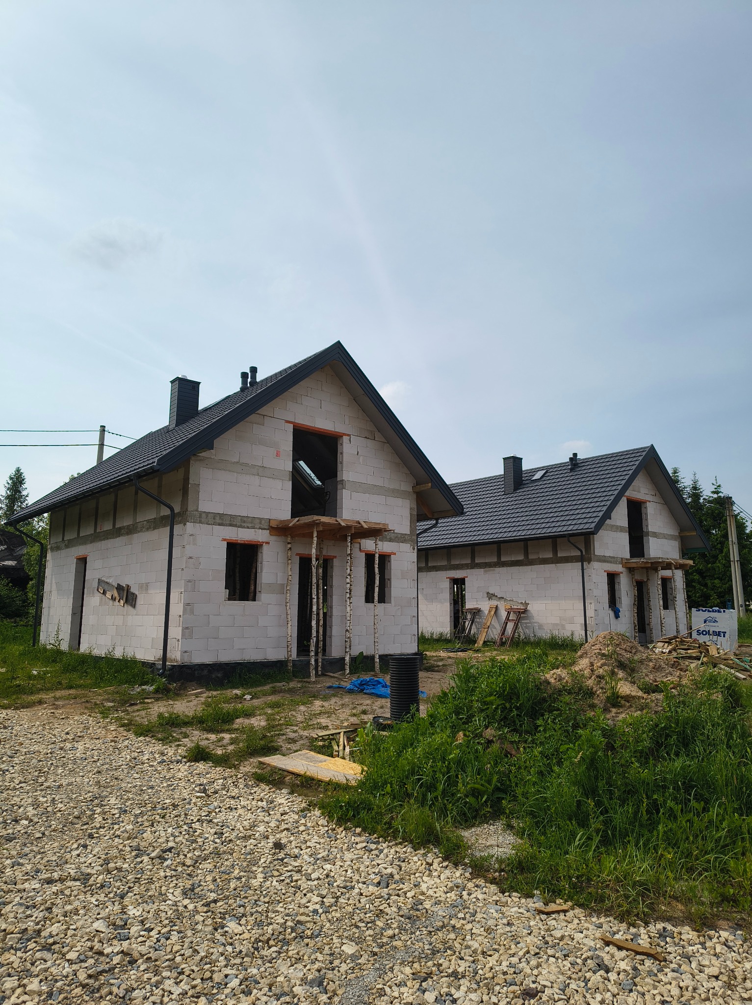 Dwa surowe, białe domy z ciemnymi dachami w trakcie budowy. Widoczne otwory okienne i drzwiowe, podparte drewnianymi stemplami. Na pierwszym planie żwirowa droga.