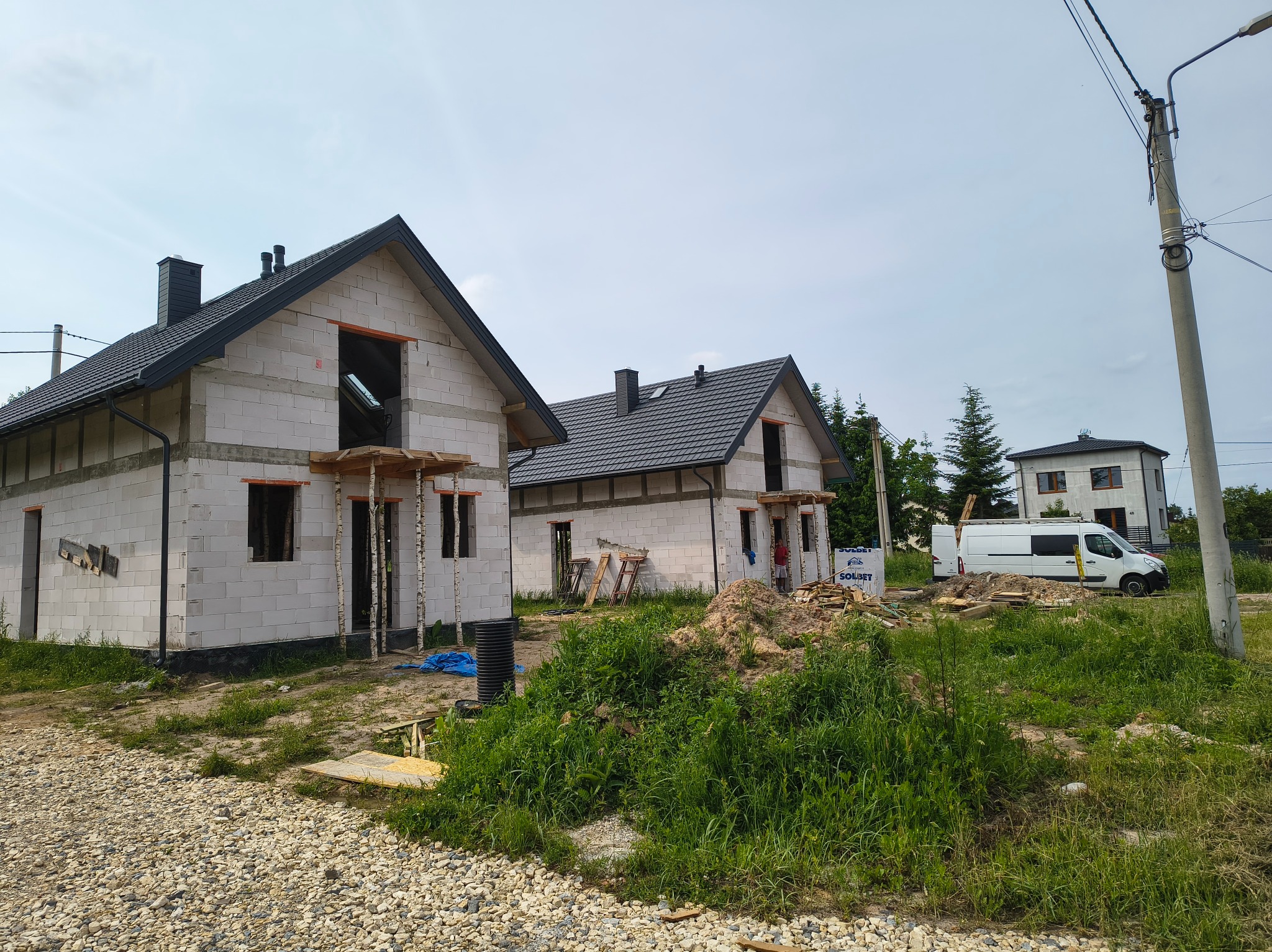 Dwa domy w stanie surowym zamkniętym z ciemnymi dachami z dachówki, białe ściany z betonu komórkowego, widoczne rynny, teren budowy z roślinnością.